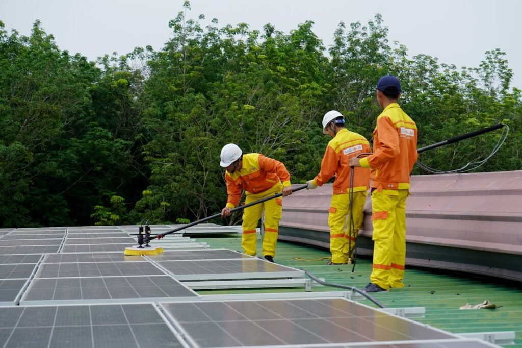 Three technicians in uniforms and hardhats clean rooftop solar panels in a renewable energy project.